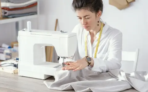 Krawcowa skupiona na pracy przy maszynie, dobiera odpowiednie rozmiary igieł do tkaniny.